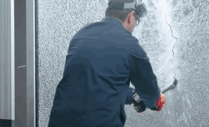 A man in protective gear strikes a large window reinforced with security window film, which shows cracking but remains intact.