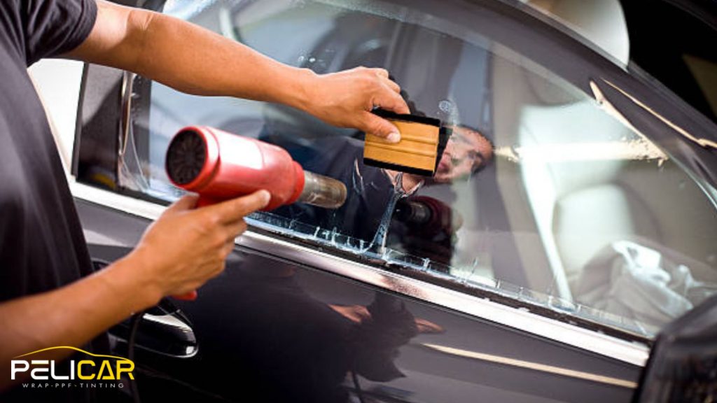 Technician using a heat gun and squeegee to apply window tint film to a car door window. The installer smooths the tint across the glass to remove bubbles and ensure a seamless professional window tint finish.