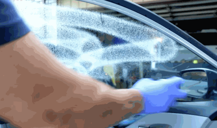 Technician wearing gloves sprays solution onto a car window to prep for window tint installation. The glass is evenly coated to help the window tint adhere smoothly and reduce imperfections.