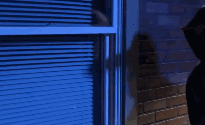 A person in a dark hoodie attempts to break into a window at night, with the glass resisting impact due to security window film.