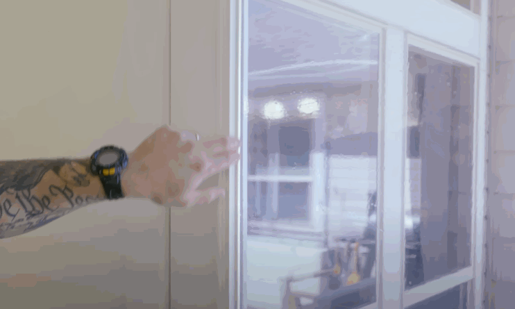 A tattooed arm wearing a black watch presses against a glass sliding door, demonstrating the installation of security window film.