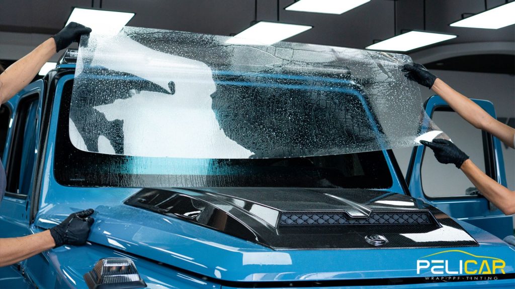 Multiple installers stretch and align a large clear sheet across the front windshield of a blue vehicle inside a workshop. The film is covered in water droplets and the text "PELICAR WRAP PPF TINTING" appears in the corner.