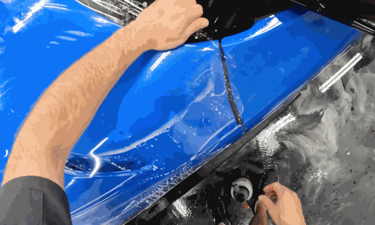 Technician stretches and smooths paint protection film over a blue car panel with water and solution visible during installation. The film conforms to the curves of the surface for a clean paint protection film finish.