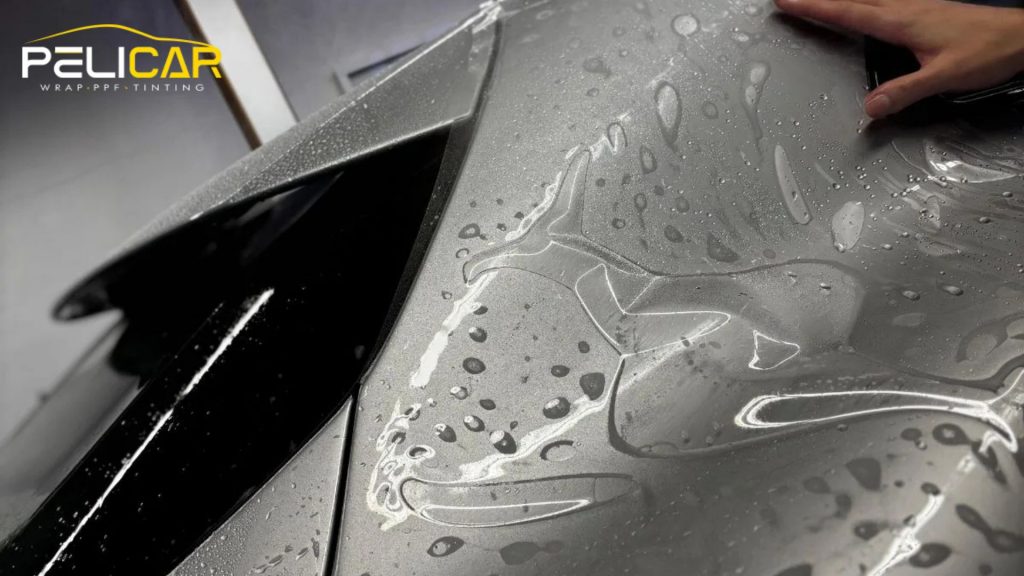 Close up of a gray car hood covered in water droplets as a technician smooths paint protection film across the surface.