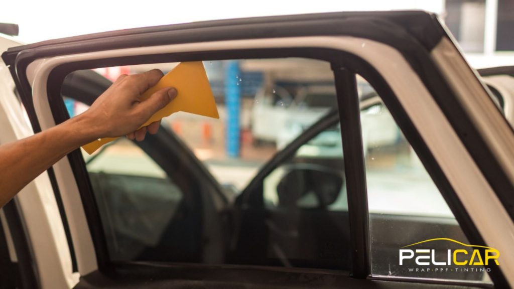 Hand using a yellow squeegee to smooth window tint film onto a car door window inside a workshop. The lower corner shows the text "PELICAR WRAP PPF TINTING" indicating professional tinting services.