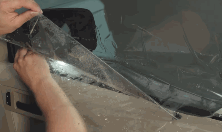 A person peels back a clear sheet of window tint film covered in water droplets while positioning it onto a car windshield. The wet surface helps adjust the film before it is secured in place.