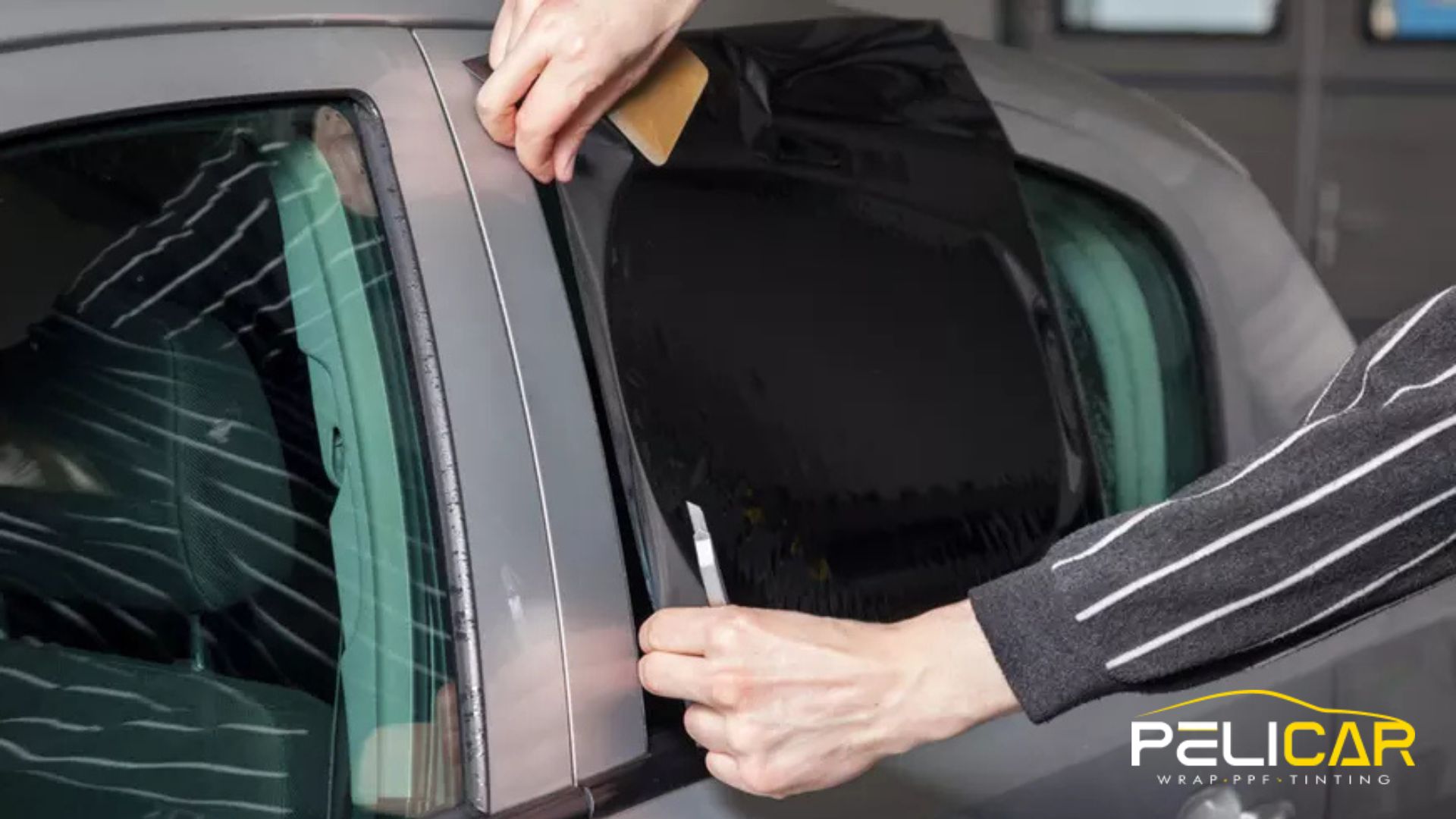 Hands apply dark window tint film to the rear side window of a gray car using a small tool to smooth it into place. The process shows careful alignment along the edge for a clean finish