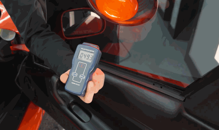 A technician holds a tint meter against a dark car window to measure visible light transmission.