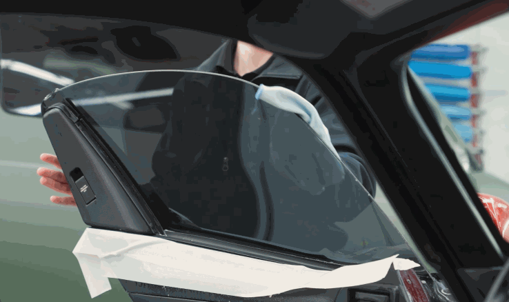 A technician positions dark window tint on an open car door window with protective tape along the lower edge. The film is being aligned before final installation.