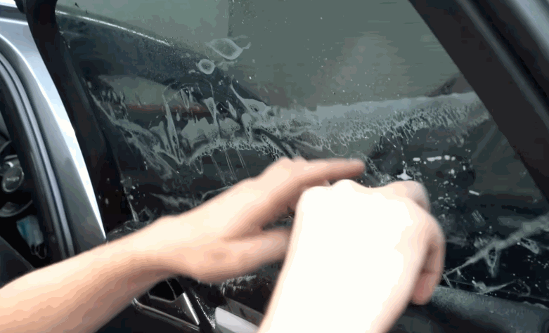 A technician wipes and smooths wet window tint on a vehicle side window. Soap and water cover the glass as the film is positioned for installation.