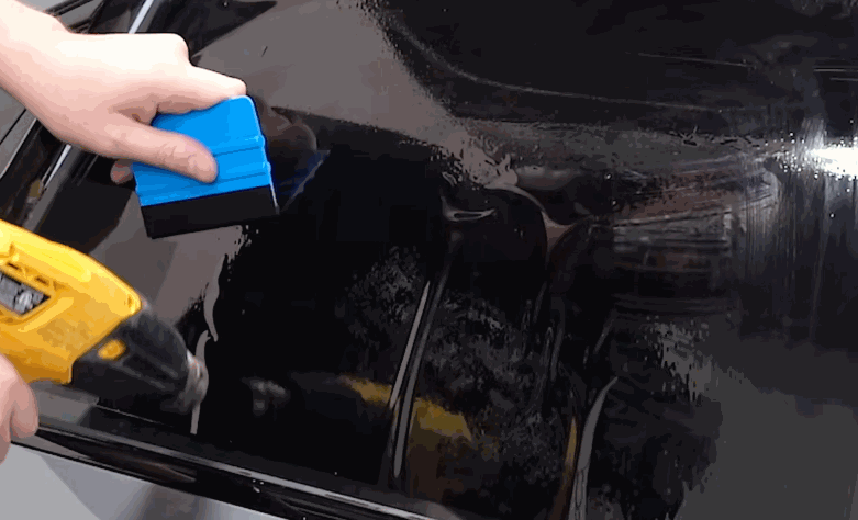 A technician uses a blue squeegee and yellow heat gun to shape window tint on a wet car window. The close up shows the film being smoothed for a clean fit.
