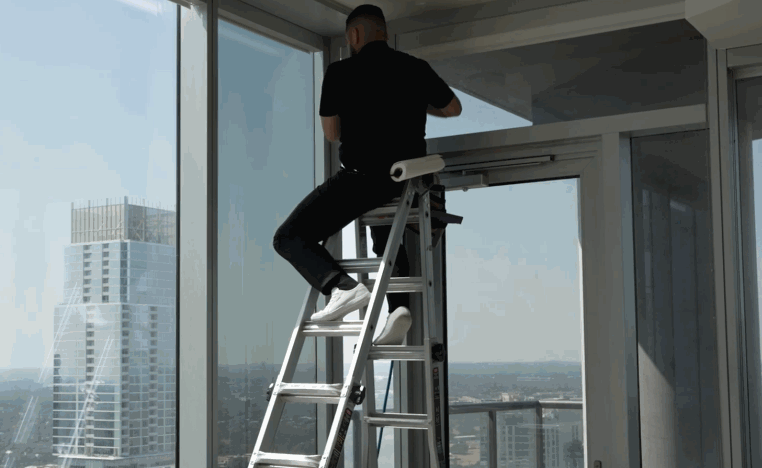 Technician on a ladder installs window film on floor-to-ceiling windows in a high-rise building.
