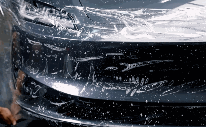 Paint protection film covers the front bumper of a black car with soap and water droplets visible across the surface.