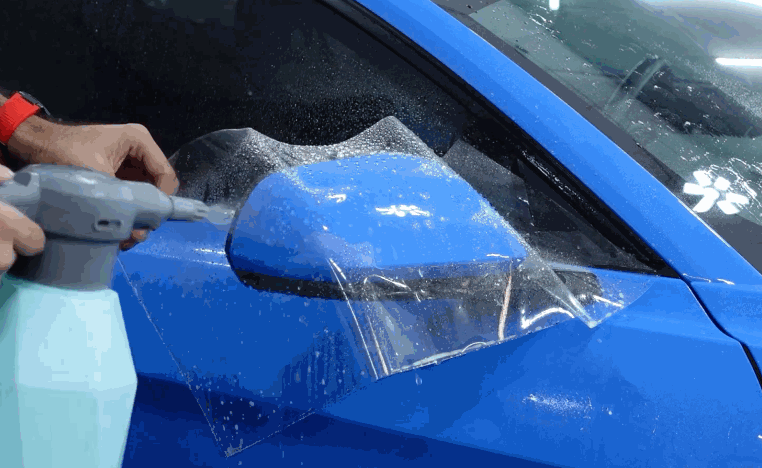 Installer sprays paint protection film over a blue side mirror to prepare it for smoothing and placement.