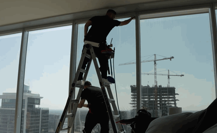 Two installers work from a ladder beside floor to ceiling office windows while applying window tint to large glass panels overlooking nearby construction.