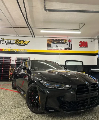 A black BMW M4 parked inside a professional window tinting shop. The car features a glossy black finish, aggressive front grille, tinted windows, and black multi-spoke wheels. The shop's branding, "peliCAR Window Tinting," is visible on the wall in the background.