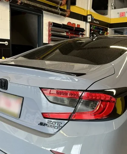 A silver Honda 2.0T Sport sedan parked inside a professional window tinting shop. The vehicle features tinted windows, black trim accents, a sporty rear spoiler, and a sleek modern design. Various tinting tools and film rolls are visible in the background.