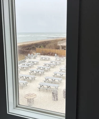 A view through a window showing an outdoor seating area with multiple picnic tables on a sandy surface near the beach. The ocean waves are visible in the background under an overcast sky.