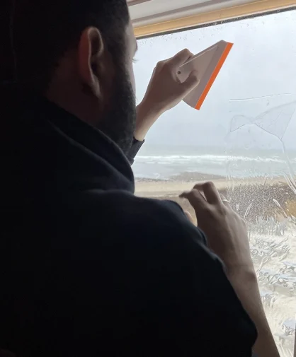 A person using a squeegee to clean a window, with a beach and parking lot visible outside.