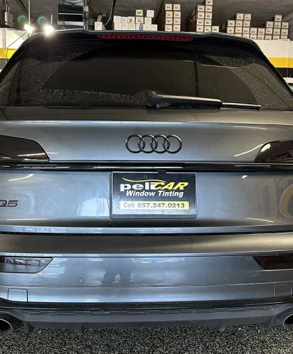 A rear view of a dark gray Audi SQ5 with tinted windows, parked inside a workshop. The license plate frame displays the Pelicar Window Tinting logo and contact number. The background includes shelves stocked with boxes and a 3M Authorized Dealer sign, indicating a professional automotive service setting.