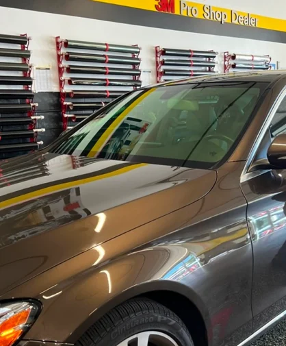 A sleek brown luxury sedan with tinted windows parked inside a professional tinting shop. The background features neatly arranged rolls of window tint film mounted on racks, along with a '3M Pro Shop Dealer' sign. The car's glossy finish reflects the workshop's interior, emphasizing its polished look.